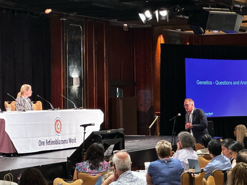 A large screen at the front of the conference room states: Genetics Q&A. A man wearing a suit asks a question at a static microphone placed among the audience, while one member of the panel is visible at the panel table, listening intently. A branded tablecloth over the panel table bears the One Rb World logo. Light reflects on the polished wooden wall paneling behind the table, creating an ethereal effect in the photo.