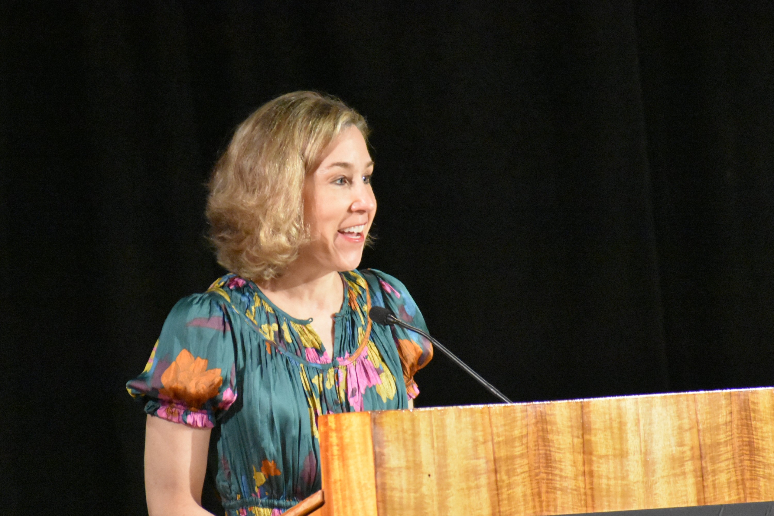 Pamela Paul stands at a light wooden podium against a black background. She wears a blue dress patterned with yellow, pink, and orange flowers.